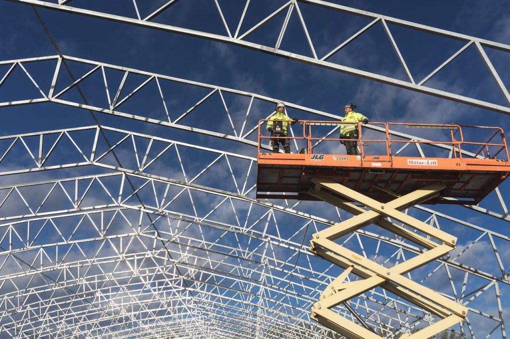 pexels-photo-13532460 Two workers on a scissor lift at a construction site, framed by a metal structure against a blue sky.